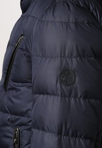 Marinblå puffjacka med kviltat mönster, utrustad med en svart rund logotyp-patch på ärmen och dragkedjefickor för funktionalitet.