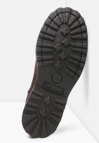 Ruskea kumisaappaan pohja, jossa on syvä kuviointi ja ristiin meneviä muotoja pidon parantamiseksi sekä kohokuvioitu logo. Tasainen, kuvioitu pinta antaa pitoa.