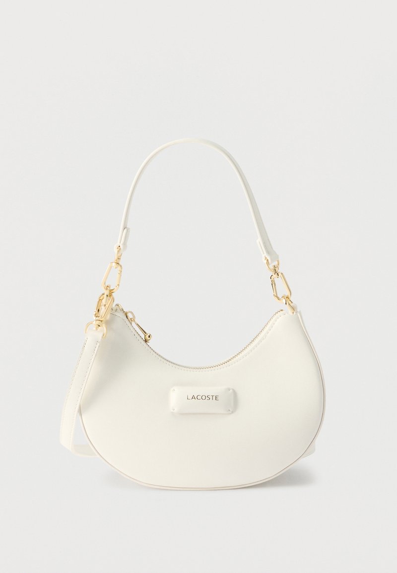 Λευκή τσάντα ώμου σε σχήμα ημισελήνου με εξαρτήματα σε χρυσή απόχρωση, κλείσιμο με φερμουάρ και λογότυπο "LACOSTE" στην μπροστινή πλευρά.