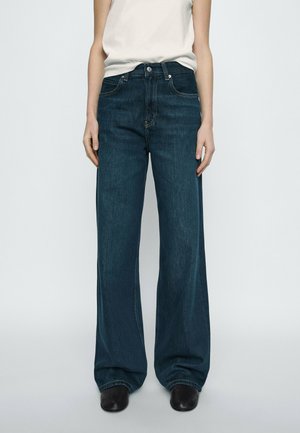 Person wearing high-waisted, wide-leg dark blue jeans with a white sleeveless top and black shoes against a plain background.