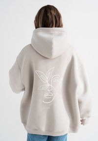 Beige hoodie met een groot geborduurd gezicht en bladdesign op de achterkant. Gemaakt van zachte stof, met een ruime capuchon en geribbelde manchetten.