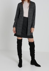Manteau long à carreaux gris, haut ajusté beige clair, jupe noir texturée et bottes en daim noir hauteur genou. Look global en couches avec des lignes épurées.