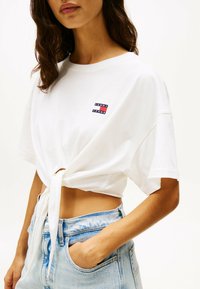 Camiseta blanca corta con mangas cortas, atada en la parte delantera, que cuenta con un pequeño parche de logo bordado en rojo, azul y negro.