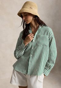 Žena v zelenej a bielej košeli s vertikálnymi pruhmi, béžovom klobúku typu bucket, bielych šortkách a náušniciach v tvare mušlí stojaca pred neutrálnym pozadím.