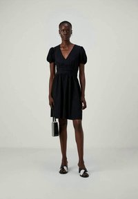 Melna īsa kleita ar V veida kakla izgriezumu, pūkainām piedurknēm un savilktu jostasvietu. Nēsāta ar mazu somiņu un slaidām sandālēm. Gluda tekstūra.