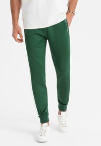 Grüne Sweatpants mit weißen Seitenstreifen, aus weichem Stoff. Verfügt über Reißverschlusstaschen und gerippte Bündchen, kombiniert mit weißen Sneakers.