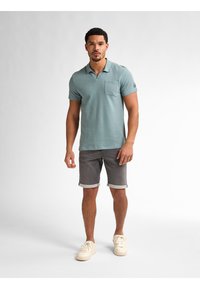Camisa de polo de manga corta en azul claro con un bolsillo, combinada con pantalones cortos grises remangados y zapatillas de deporte color crema. Diseño sencillo, corte casual.