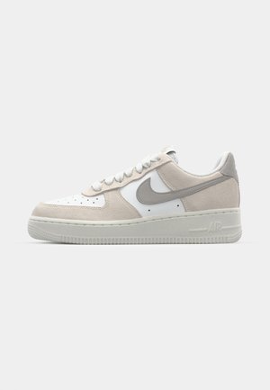 Sneakers Nike Air Force 1 avec des panneaux en cuir blanc et en daim beige, des lacets ronds, des accents gris et une semelle en caoutchouc texturée.