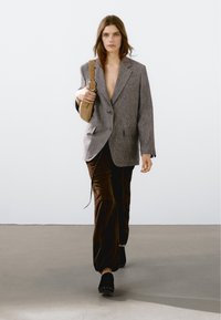 Donna che cammina in avanti indossando un blazer grigio oversize, pantaloni in velluto marrone, scarpe nere e portando una borsa a tracolla beige su uno sfondo semplice.