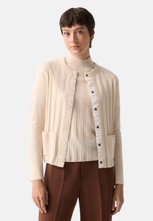 Ung kvinde med kort mørkt hår iført en cremefarvet ribstrikket cardigan over en matchende rullekravesweater og brune bukser, stående mod en hvid baggrund.