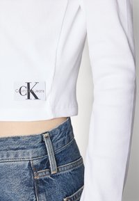 Balta šonkaine iškirpta viršutinė dalis su ilgomis rankovėmis, puikuojasi Calvin Klein Jeans etikete. Derinama su vidutinio plovimo džinsais, kuriuose matomi siūlai.