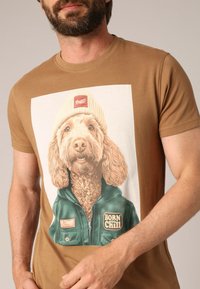 Bruine katoenen t-shirt met een afbeelding van een hond die een beige beanie en groene jas draagt, met de tekst "BORN TO CHILL" en details.