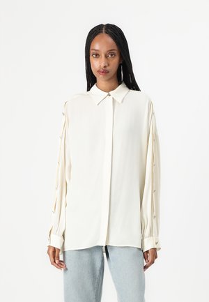 Blusa crema con colletto, maniche lunghe con dettagli a bottoni e una texture liscia. Abbinata a jeans blu chiaro.