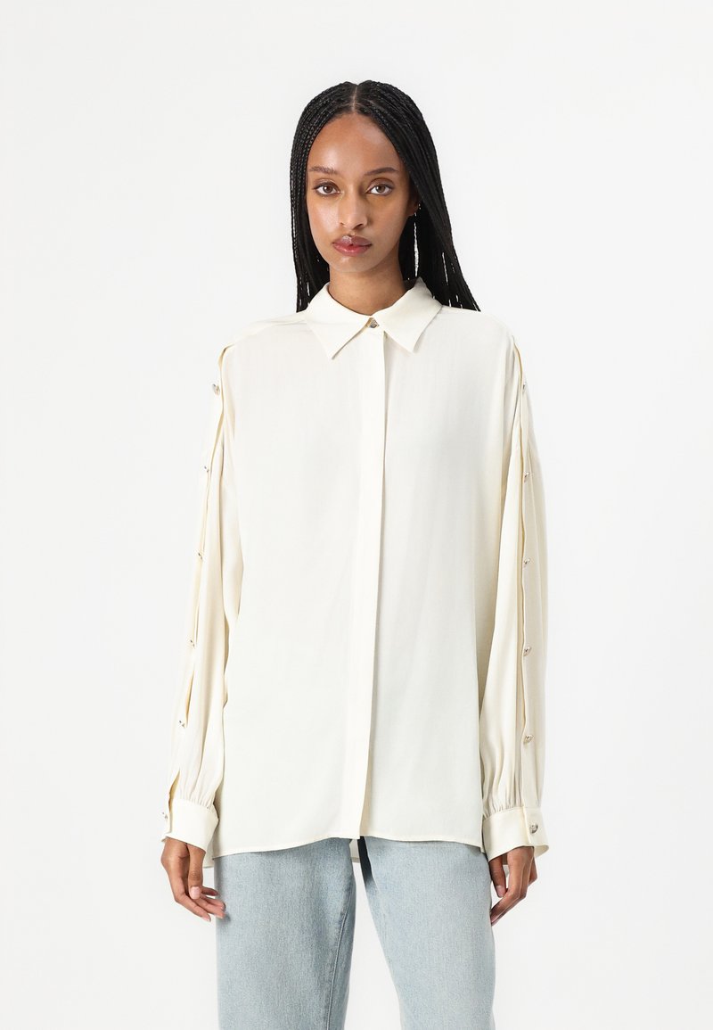 Blusa crema con colletto, maniche lunghe con dettagli a bottoni e una texture liscia. Abbinata a jeans blu chiaro.