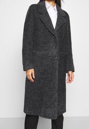 Femme portant un long manteau texturé gris foncé avec deux boutons sur une chemise blanche et un pantalon noir, debout devant un fond uni.
