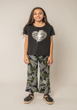 Jeune fille debout et souriante, portant un t-shirt noir avec un cœur et un éclair, ainsi qu'un pantalon large à motifs de jungle.