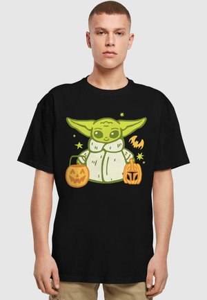 Black t-shirt featuring a green character holding two orange Halloween pumpkins, with a playful design and simple background shapes.