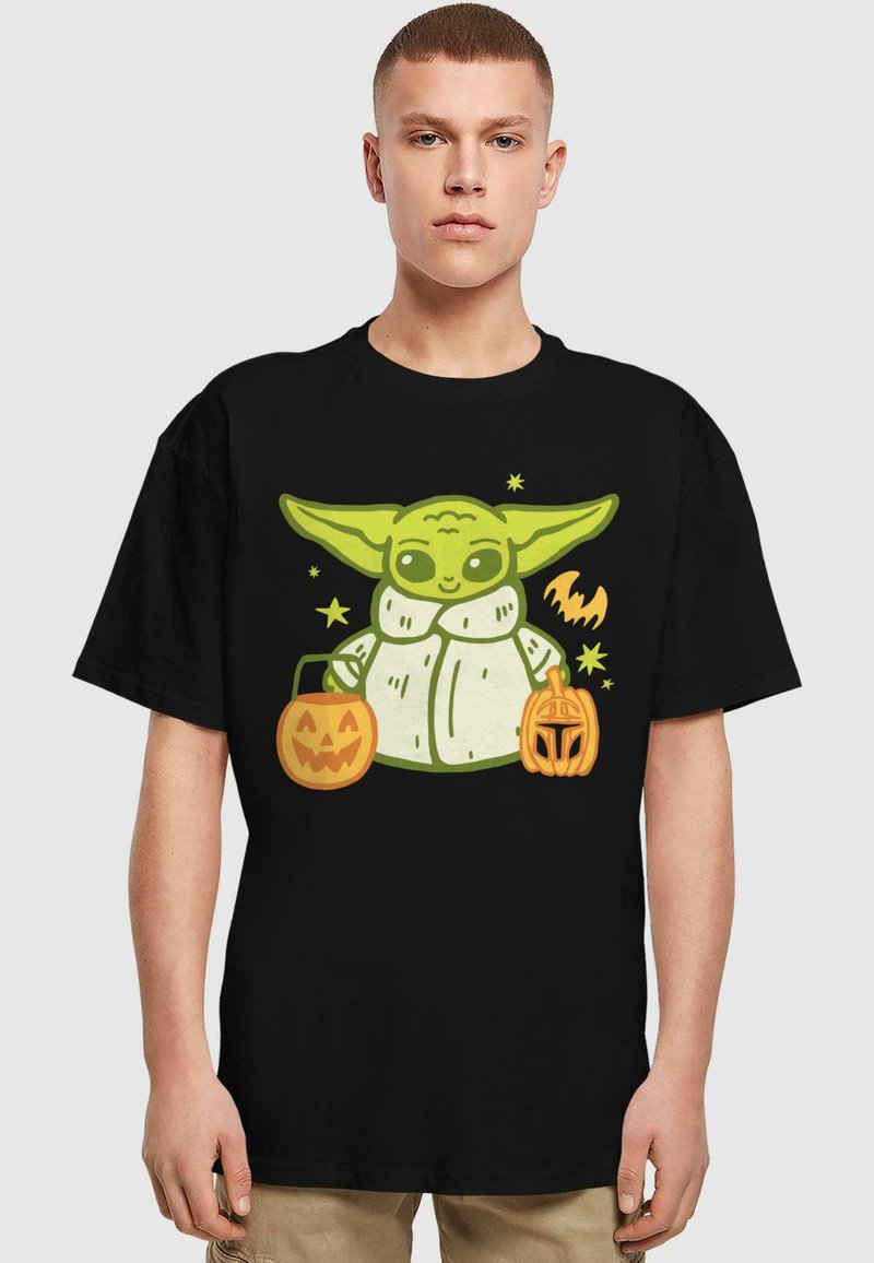 Čierne tričko s obrázkom zelenej postavy, ktorá drží dve oranžové tekvice na Halloween, s hravým dizajnom a jednoduchými pozadím tvarmi.