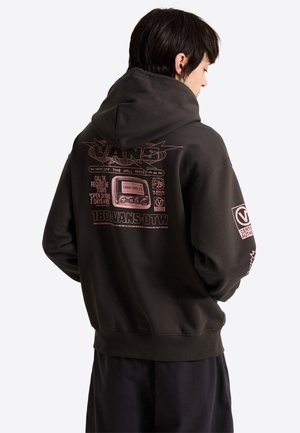 Person trägt schwarzen Hoodie mit pinkem grafischem Text und Vintage-Radio-Design auf dem Rücken, steht mit dem Rücken zu einem schlichten weißen Hintergrund.