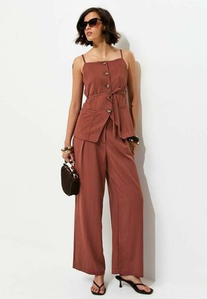 Femme portant un haut sans manches boutonné couleur rouille avec ceinture à nouer à la taille et un pantalon large assorti, tenant un sac à main noir et portant des lunettes de soleil.