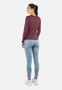 Frau mit Pferdeschwanz, die goldene Ohrringe, ein langärmliges weinrotes Oberteil, helle Jeans und weiße Sneakers trägt, steht und zur Seite schaut.