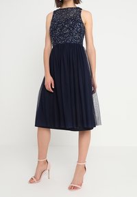 Robe sans manches bleu marine avec un corsage en sequins et une jupe en tulle fluide, présentant un design à hauteur de genoux et des sandales à talons hauts rose pâle.
