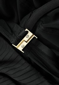 Juoda medžiaga su šiek tiek tekstūruota paviršiaus struktūra, puošta auksiniu metaliniu sagtimi su dviem varžtais, esančia persidengiančių dirželių dizaine.