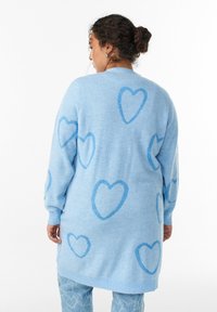 Svetlomodrý cardigan s jemnou textúrou, zdobený vzorom tmavomodrých srdiečok. Dlhé rukávy a uvoľnený strih.