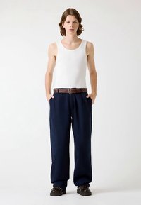 Débardeur blanc, pantalon large bleu marine avec ceinture, chaussures foncées. Le mannequin se tient les mains dans les poches devant un fond clair.