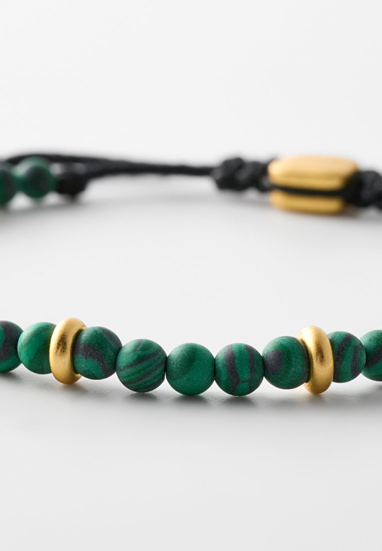 Perles marbrées vertes et noires avec des accents en métal doré, disposées sur un bracelet en cordon noir. Fermoir ajustable avec perle dorée.