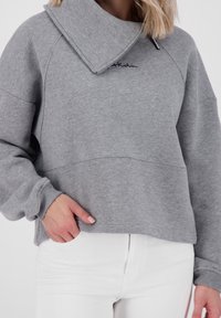 Grauer Pullover mit weitem, umgeschlagenem Kragen, lockerer Passform und gerippten Bündchen. Verfügt über Logo-Stickerei und strukturierte Materialien. Kombiniert mit weißen Jeans.