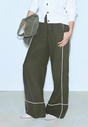 Person iført olivengrønne bukser med vide ben og hvid piping, holder en matchende grøn taske, hvid skjorte og hvide mesh-sko.