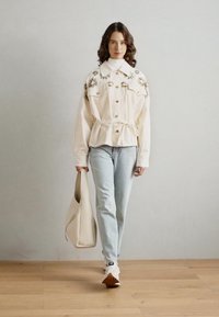 Giacca di jeans crema con ricami floreali, vita cinchata, abbinata a jeans azzurri chiari e sneakers bianche, mentre tiene una grande borsa tote crema.