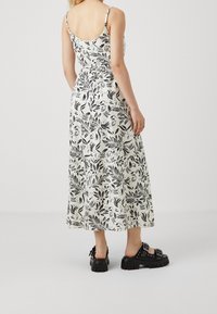 Robe maxi blanche à motif floral avec des feuilles noires, bretelles fines et taille cintrée. Associée à des sandales noires épaisses.