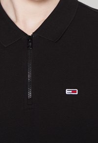 Svart polo-shirt med ett strukturerat tyg, med en dragkedja fram och en liten broderad logotyp i röd, vit och blå på vänster bröst.