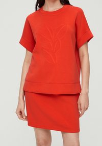 Top de mezcla de algodón rojo con manga corta, que presenta un diseño en relieve de hojas y un dobladillo recto. Combinado con una falda roja a juego.