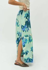 Gonna maxi in tessuto verde chiaro con motivi astratti blu, dotata di uno spacco laterale. Indossata con scarpe slip-on grigie, che mostrano un tacco piatto.