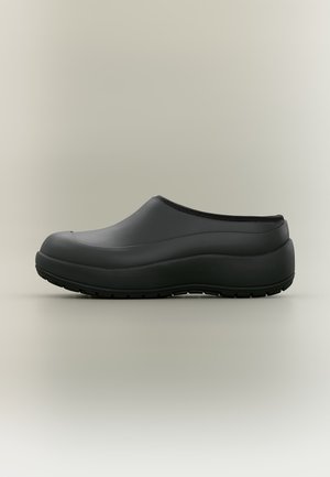 Zueco negro sin cordones con suela gruesa y punta redondeada, diseñado para un fácil uso y para actividades al aire libre sobre un fondo neutro.
