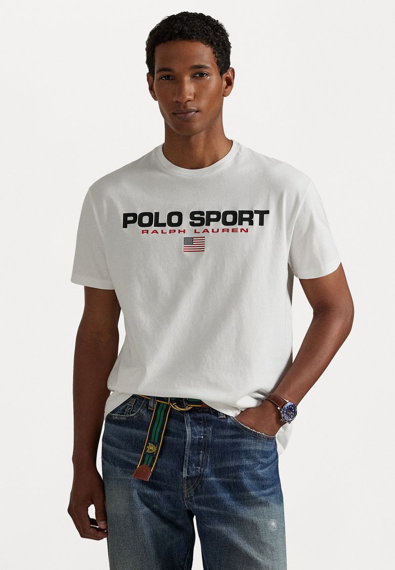 Tricou alb din bumbac, cu text bine definit "POLO SPORT RALPH LAUREN" în negru și roșu, detaliu cu steagul american, guler rotund și mâneci scurte.