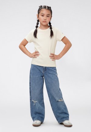 Jong meisje met gevlochten haar staat met handen in de zij, draagt een crèmekleurig T-shirt, gescheurde losse blauwe spijkerbroek en witte sneakers tegen een effen achtergrond.