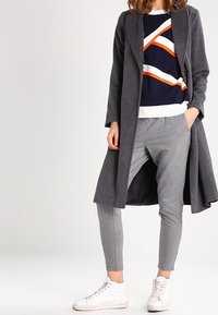 Grå långjacka över en marinblå och vit randig tröja med orange accenter, matchat med grå joggers och vita sneakers.