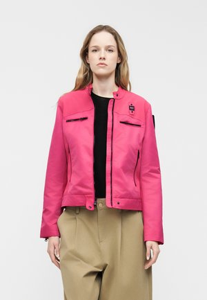 Femme portant une veste zippée rose vif sur un haut noir et un pantalon beige, debout devant un fond clair uni.