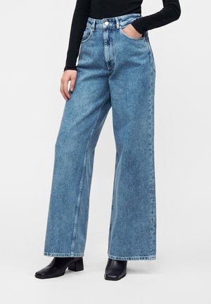 Personne portant un jean bleu taille haute à jambes larges, des bottines noires et un haut noir à manches longues, debout avec une main dans la poche.