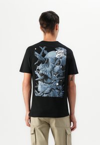 Zwart katoenen t-shirt met een blauwe schedelafbeelding op de achterkant, bestaande uit hoekige, gebroken vormen en abstracte witte accenten. Korte mouwen.