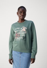 Zelená mikina s kulatým výstřihem, s grafikou v růžové a černé barvě s textem "Život je lepší mezi stromy." Jednoduchý design, fleecový materiál.