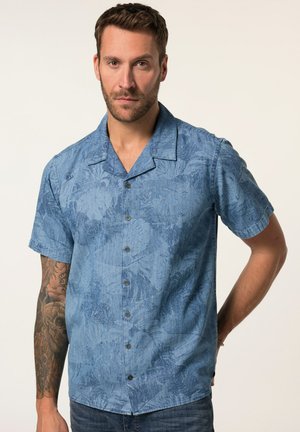 Uomo con braccio destro tatuato che indossa una camicia blu a maniche corte con un delicato motivo a foglie, in piedi con la mano sinistra dietro la schiena.