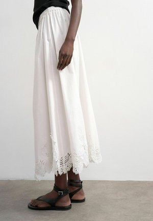 Jupe longue blanche avec bordure en dentelle à l'ourlet, portée avec des sandales plates noires à brides et un haut noir, debout sur un sol gris contre un mur blanc.