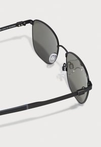 Ochelari de soare negri, cu lentile gri, având o formă geometrică unică, cu garnituri de nas transparente și pârghii subțiri negre.