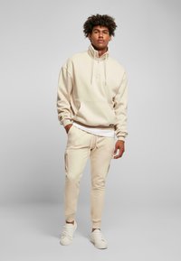 Beige hette med halv glidelås i kragen, paired med matchende joggebukser med side lommer og ribbestrikkede mansjetter. Brukt med hvite joggesko.
