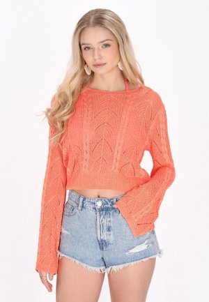 Pull en crochet court de couleur corail vif avec un motif de feuilles texturé. Associé à un short en jean clair déchiré.